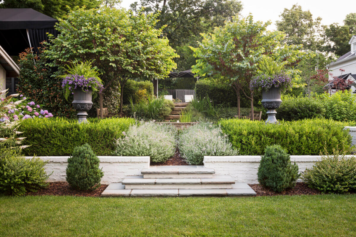 Daniel Keeley on Designing Outdoor Spaces That Actually Get Used - 3 A landscaped garden by Daniel Keeley with stone steps, trimmed bushes, white brick walls, potted plants, and trees exudes a sophisticated style, leading to a serene seating area in the background.