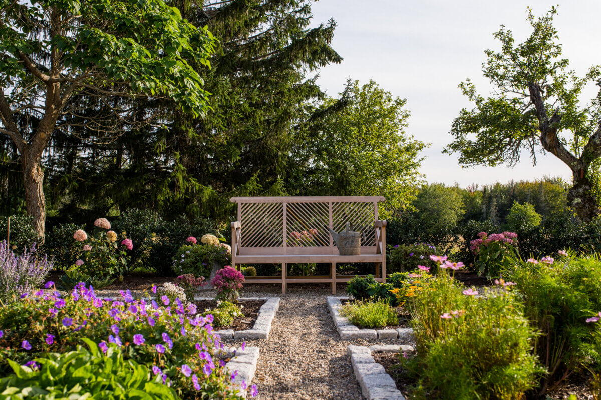 Daniel Keeley on Designing Outdoor Spaces That Actually Get Used - 5 A wooden bench, designed in a sophisticated style by Daniel Keeley, sits at the end of a gravel path in a garden, surrounded by blooming flowers, green shrubs, and trees on a sunny day.
