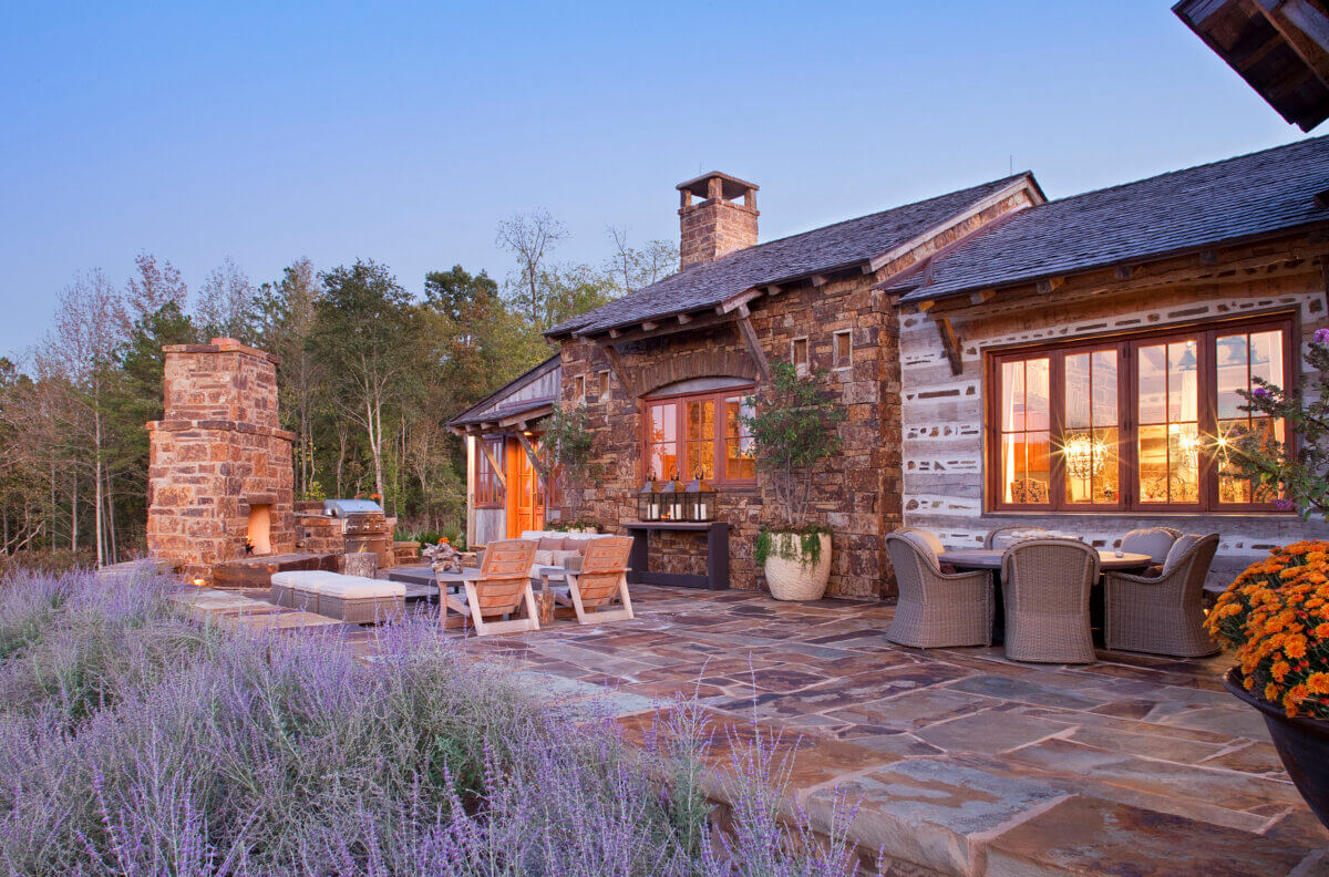 Daniel Keeley on Designing Outdoor Spaces That Actually Get Used - 7 Stone patio with outdoor seating, dining area, and fireplace, bordered by lavender plants, showcases sophisticated style in front of a rustic stone and wood house designed by Daniel Keeley at sunset.