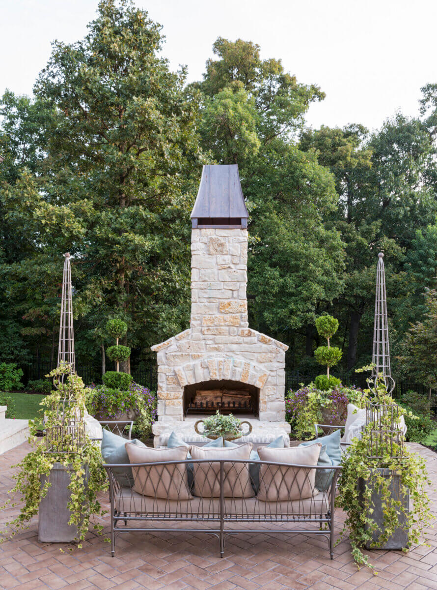 Daniel Keeley on Designing Outdoor Spaces That Actually Get Used - 9 Outdoor patio with a stone fireplace, metal sofa with cushions, potted plants, and trees in the backgroundβshowcasing Daniel Keeleyβs sophisticated style.
