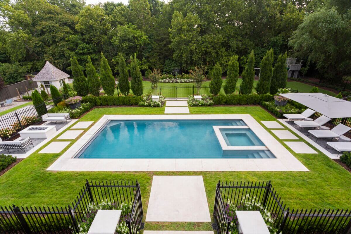 Daniel Keeley on Designing Outdoor Spaces That Actually Get Used - 10 A rectangular backyard pool with an attached square hot tub, designed by Daniel Keeley, exudes sophisticated style; it's surrounded by lounge chairs, manicured lawn, and tall trees in the background.