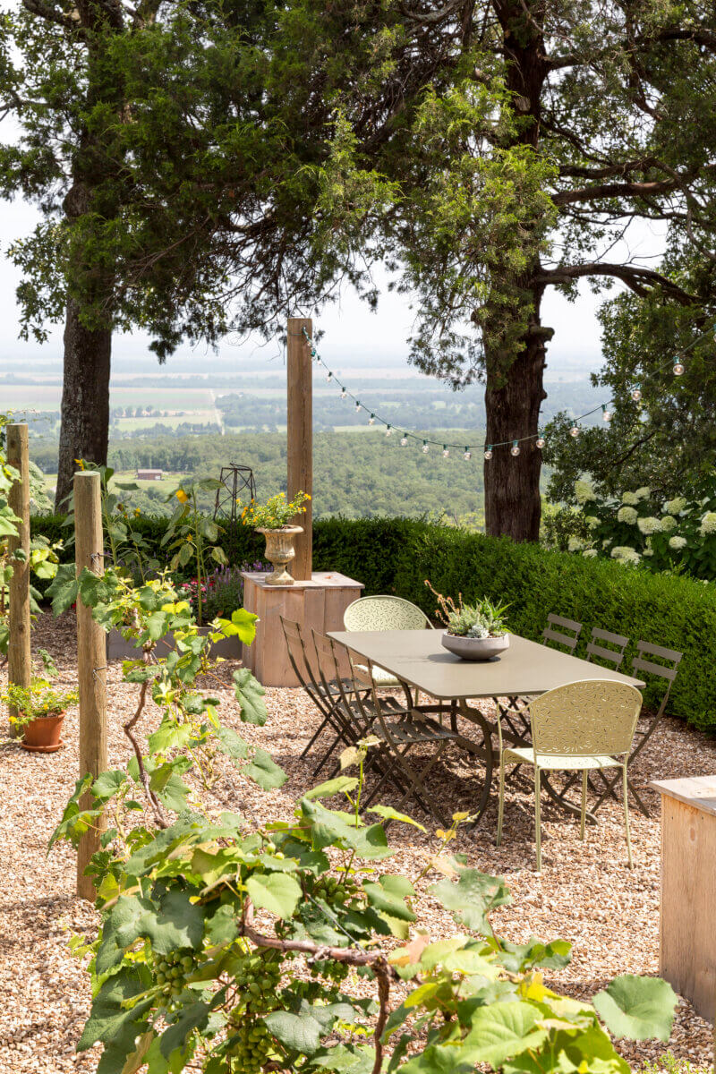 Daniel Keeley on Designing Outdoor Spaces That Actually Get Used - 14 Outdoor dining area with a long table and chairs set on gravel, surrounded by greenery and trees, featuring string lights and a scenic viewβan inviting space that reflects Daniel Keeley's sophisticated style.