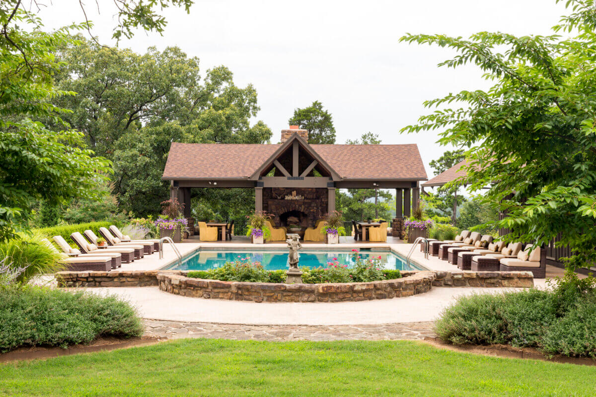 Daniel Keeley on Designing Outdoor Spaces That Actually Get Used - 12 Rectangular swimming pool with lounge chairs surrounds a stone patio, a small fountain in front, and a covered outdoor fireplace area in the background, showcasing Daniel Keeleyβs sophisticated style amid lush trees and greenery.