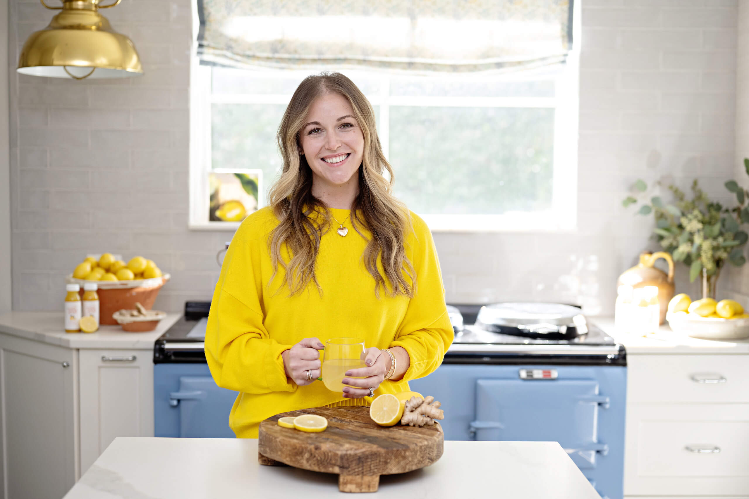 A woman in a yellow sweater stands in a bright kitchen, holding a mug near a cutting board with lemon slices and ginger. Lemons and bottles are visible on the counter behind her.