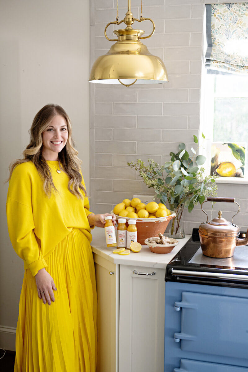 Trusting Her Gut: Meet the Charlotte Woman Making Healthy Habits Easier - 7 A woman in a bright yellow dress stands in a kitchen next to a bowl of lemons, ginger, bottled drinks, and a copper kettle on a blue stove.