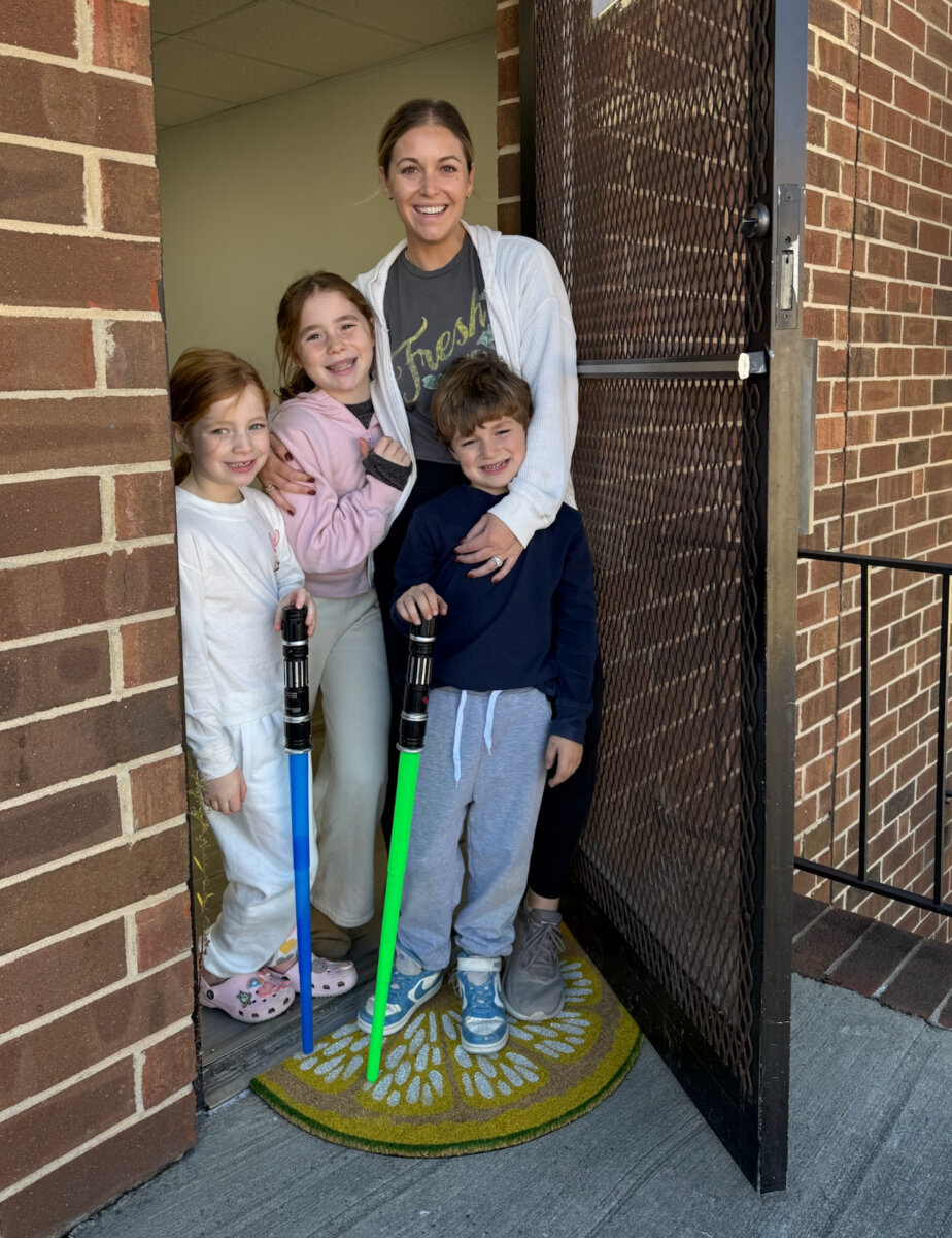 Trusting Her Gut: Meet the Charlotte Woman Making Healthy Habits Easier - 6 A mom entrepreneur and her three children stand at an open door on a sunflower doormat. Two children hold toy lightsabers, one green and one blue. The group is smiling.