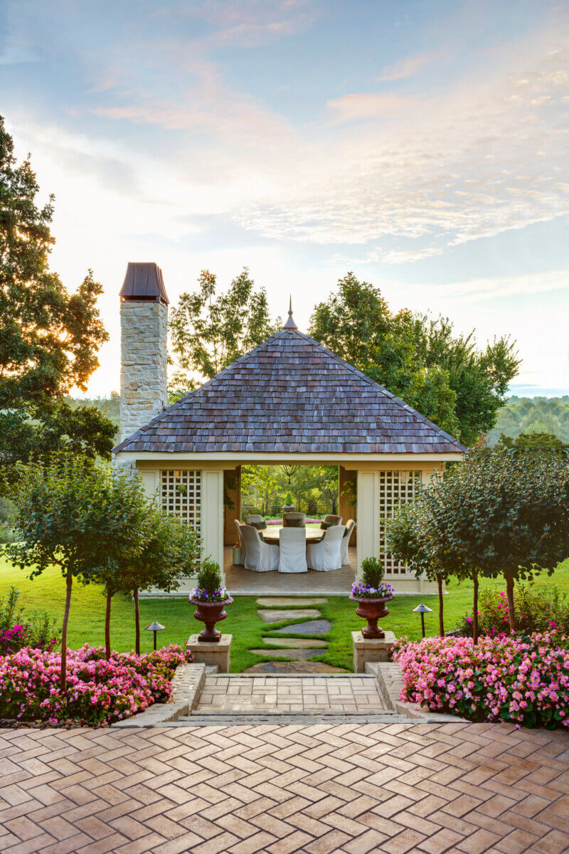 Daniel Keeley on Designing Outdoor Spaces That Actually Get Used - 6 A garden gazebo by Daniel Keeley, featuring a shingled roof, stone chimney, and outdoor dining area, exudes sophisticated style amid green grass, pink flowers, and trees beneath a partly cloudy sky.