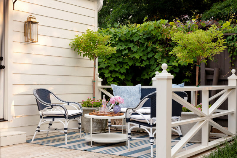 Daniel Keeley on Designing Outdoor Spaces That Actually Get Used - 4 A small outdoor patio with sophisticated style by Daniel Keeley features two chairs, a low table, blue and white cushions, potted plants, and a striped rug next to a cream-colored house.