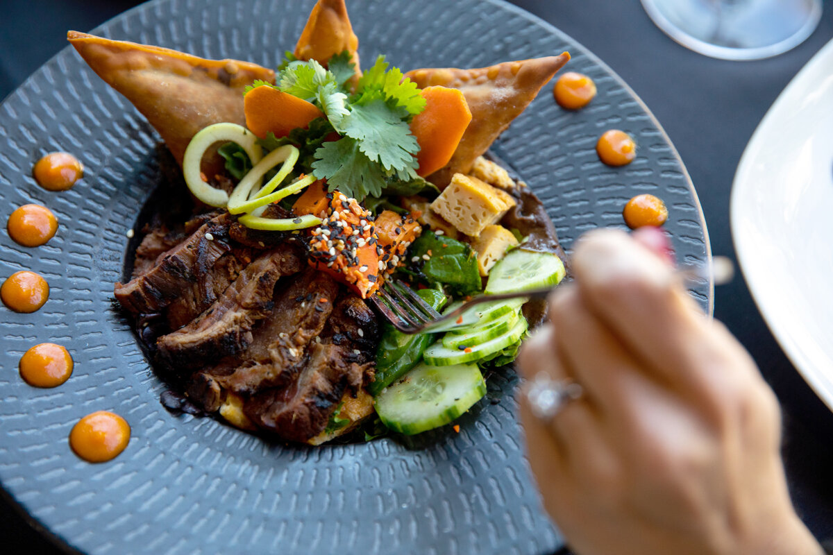 A plate of sliced grilled meat with fresh vegetables, herbs, sauce dots, and two fried triangles; a hand holds a fork near the food—perfect for anyone searching for the best lunch Nashville has to offer.