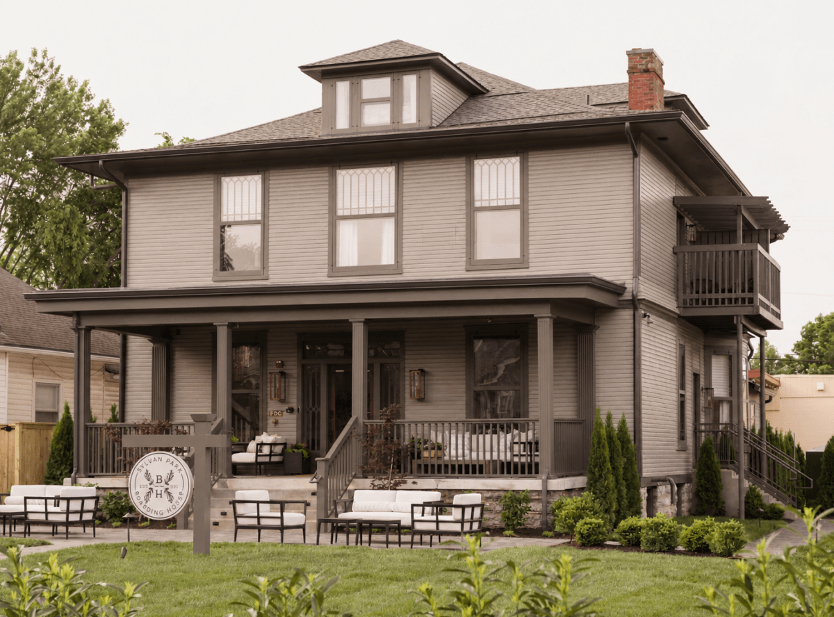 Sylvan Park Boarding House features a covered front porch with outdoor seating, a small balcony, and a sign in the yard. Shrubs and grass surround the inviting home.