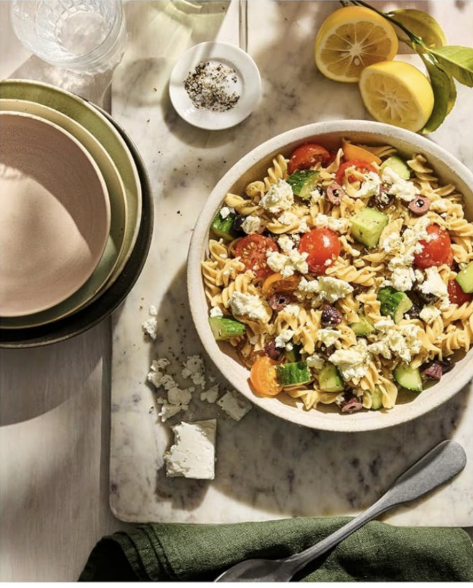 RECIPE: Chickpea Pasta Salad - 2 A bowl of pasta salad with cherry tomatoes, cucumber, olives, and feta cheese sits on a marble surface, beside lemon halves, stacked bowls, and a small dish of pepper.