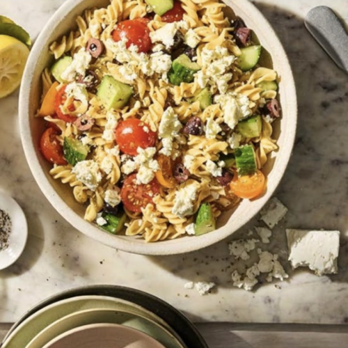 RECIPE: Chickpea Pasta Salad - 3 A bowl of pasta salad with cherry tomatoes, cucumbers, olives, and crumbled feta cheese sits on a marble surface beside lemon halves, a fork, a knife, and a cloth napkin.
