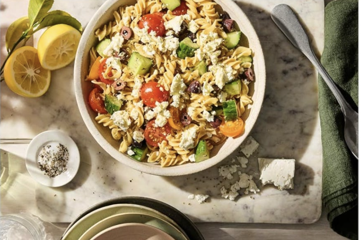 A bowl of pasta salad with cherry tomatoes, cucumbers, olives, and crumbled feta cheese sits on a marble surface beside lemon halves, a fork, a knife, and a cloth napkin.