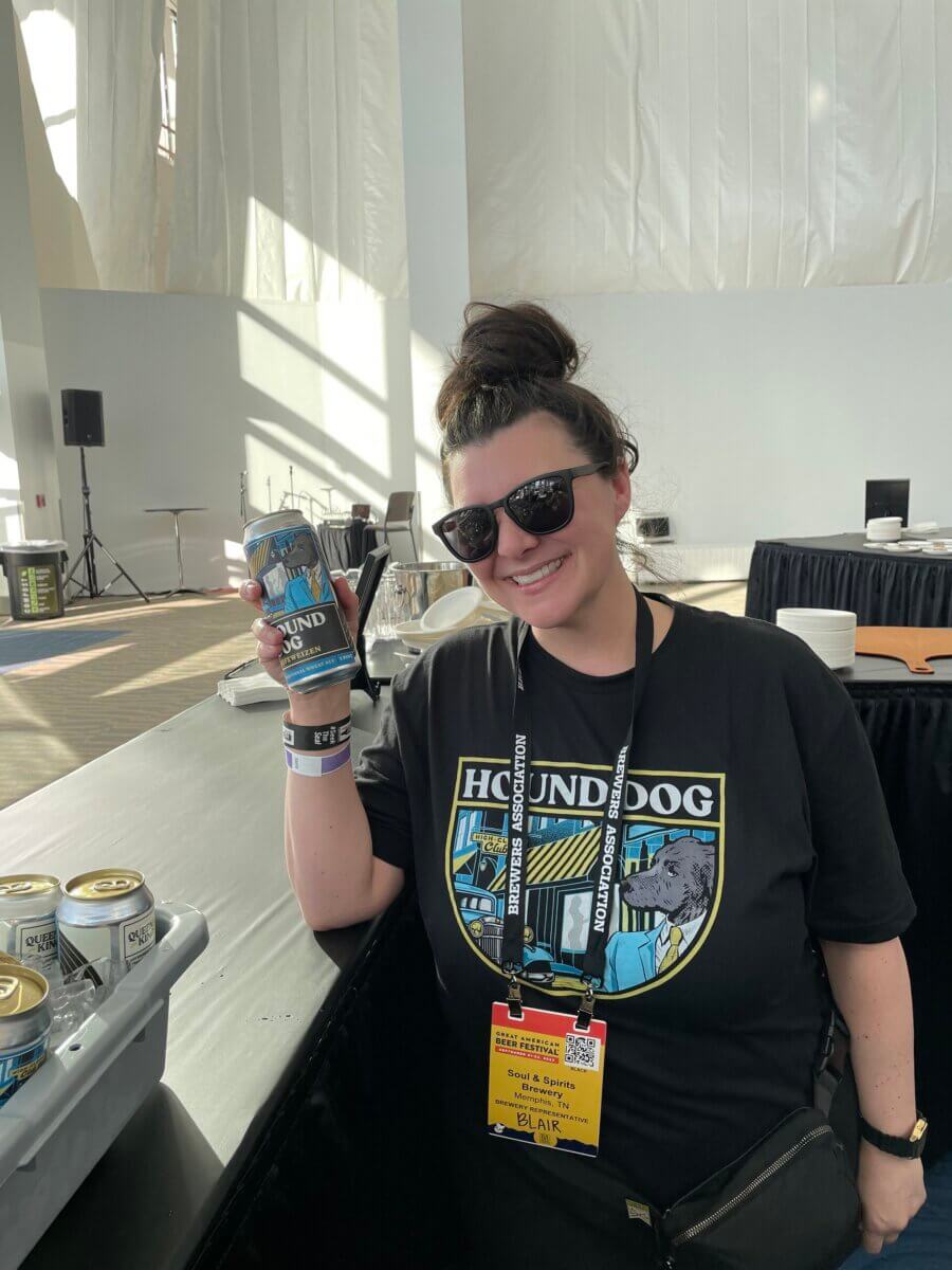 A woman wearing sunglasses and a Hounddog t-shirt stands indoors, smiling and holding a can of Hounddog beer. Event lanyards and a name badge are around her neck.