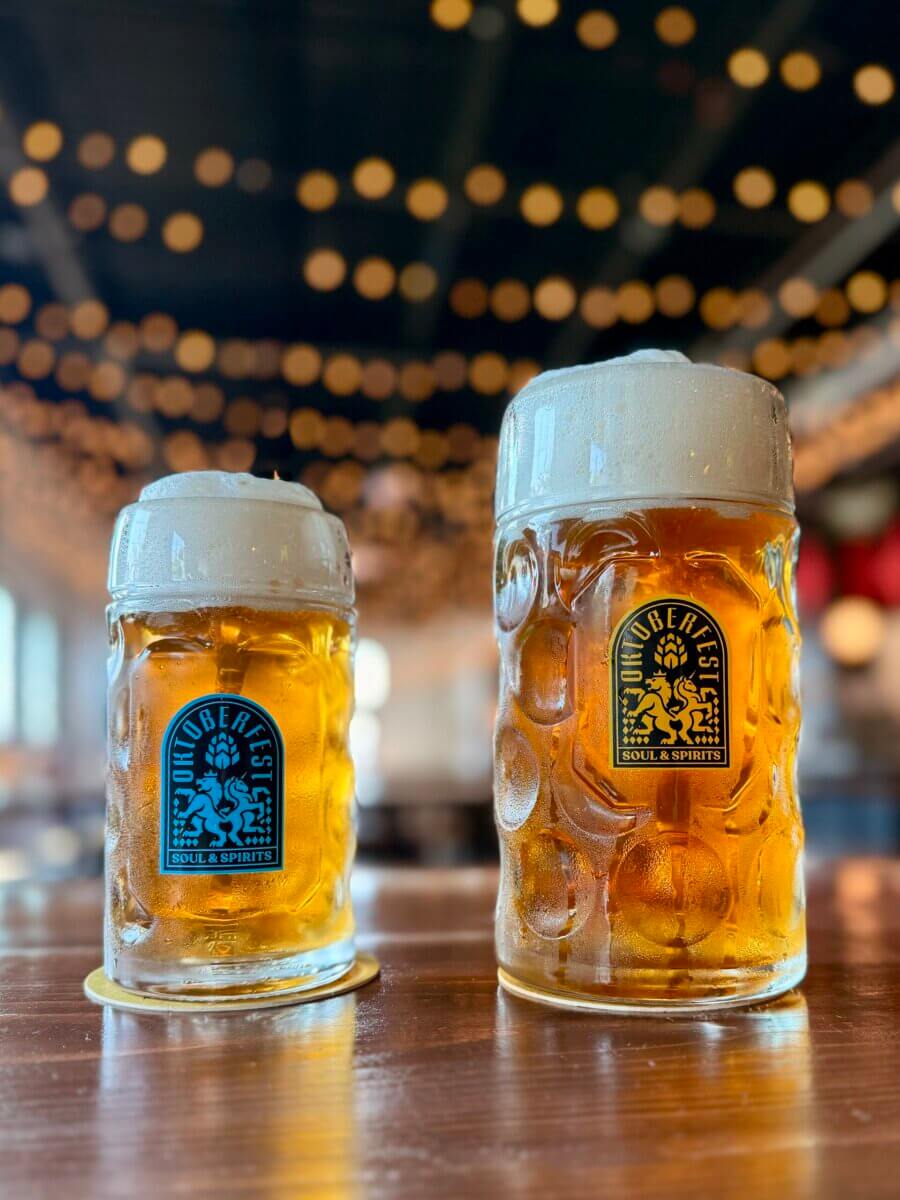 Two glass mugs of beer with foam on top, labeled with "Noble Fest" logos, sit on a wooden table with string lights blurred in the background.