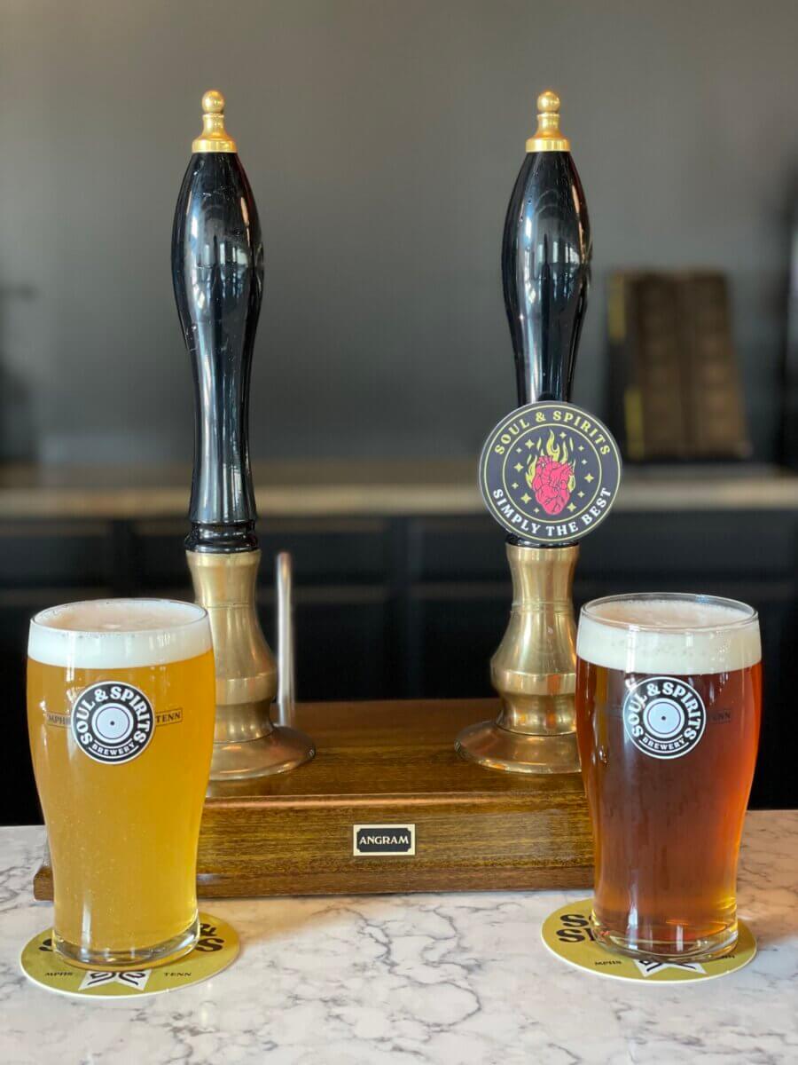 Two pint glasses of beer, one light and one amber, sit on coasters in front of two traditional hand-pull beer pumps on a marble counter.