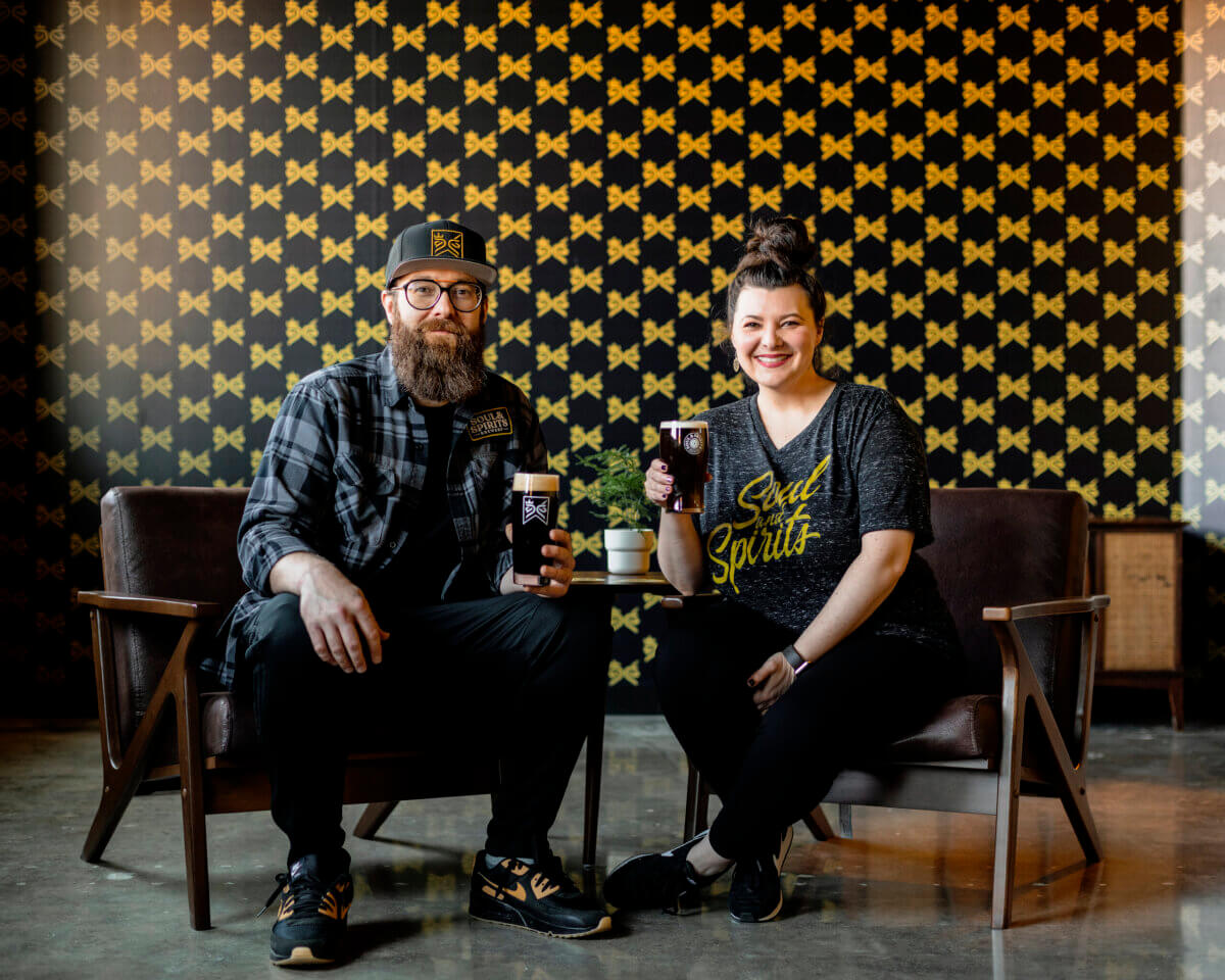 Two people sit in chairs holding drinks, smiling at the camera, with a patterned black and gold wall in the background.
