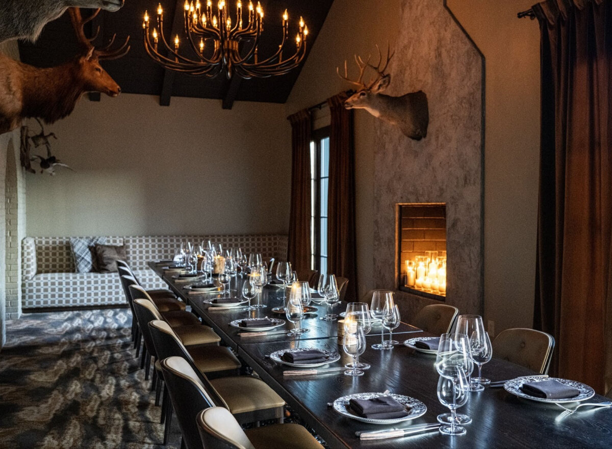 Your Guide to Sylvan Park: Nashville's Coziest Neighborhood - 3 A long dining table set with plates and glassware sits in a dimly lit Sylvan Park room, with a lit fireplace, chandeliers, and mounted deer heads guiding the rustic ambiance.