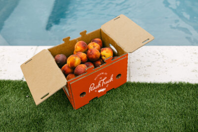 A cardboard box labeled "Peach Truck" filled with peaches sits on artificial grass beside a swimming pool, perfect for celebrating Peach Week in Nashville with fresh peach dishes.
