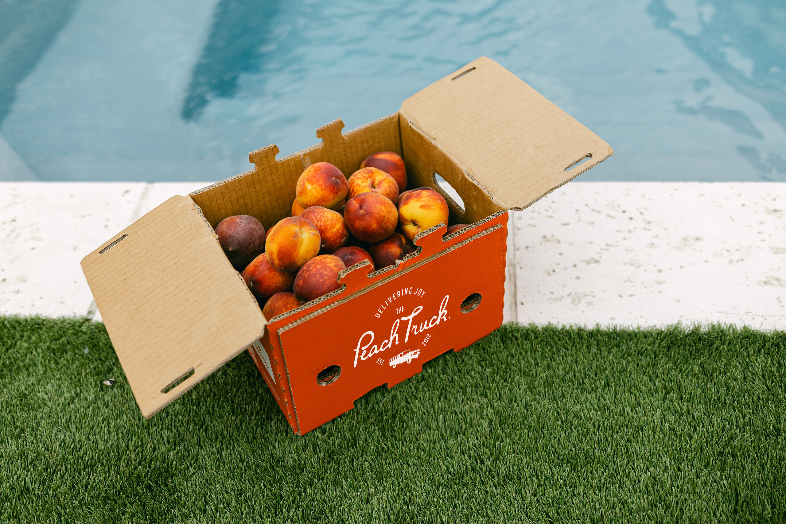 A cardboard box labeled "Peach Truck" filled with peaches sits on artificial grass beside a swimming pool, perfect for celebrating Peach Week in Nashville with fresh peach dishes.
