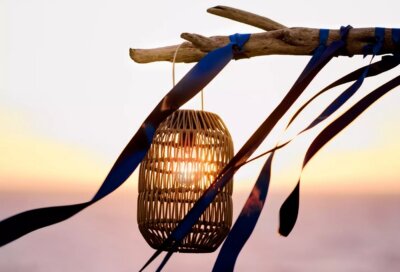 A wicker lantern hangs from a driftwood branch with blue ribbons, illuminated by sunlight, against a blurred sunset sky—perfect for outdoor entertaining.