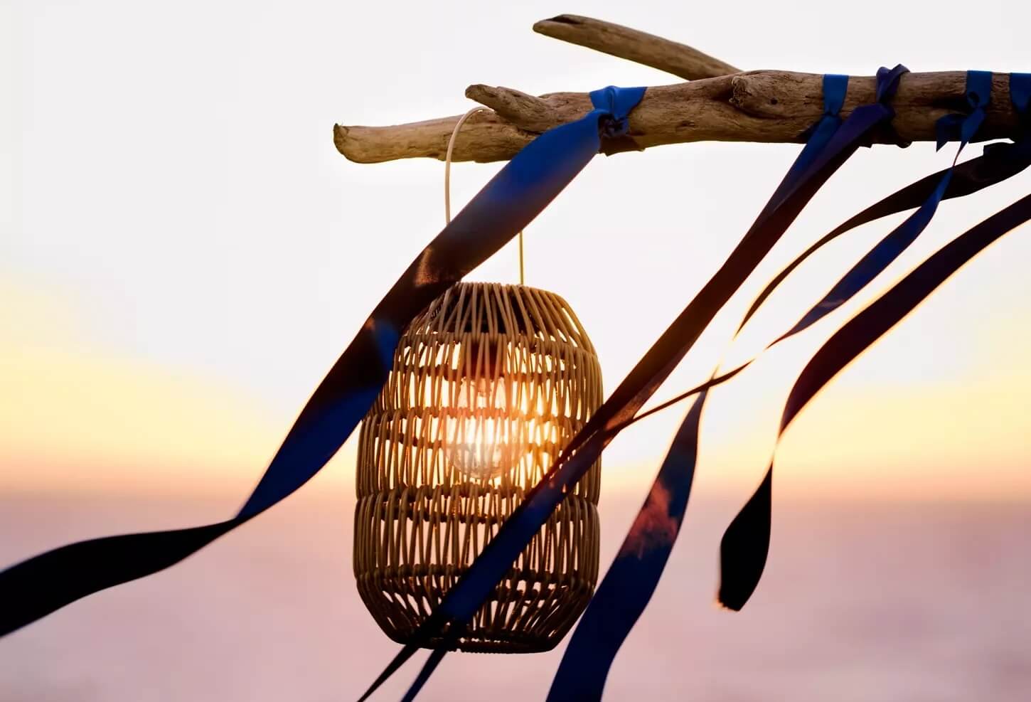 A wicker lantern hangs from a driftwood branch with blue ribbons, illuminated by sunlight, against a blurred sunset sky—perfect for outdoor entertaining.