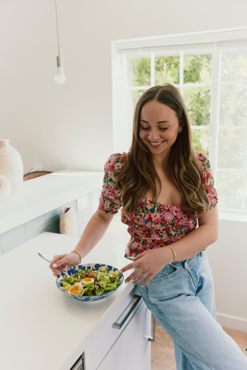 A woman stands in a bright kitchen, smiling and holding a fork over a bowl of salad with sliced boiled eggs, prepping hormone balancing foods to support cycle syncing.