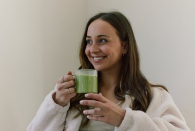 A woman with long brown hair smiles while holding a clear mug filled with a green beverage, standing against a plain light-colored background.