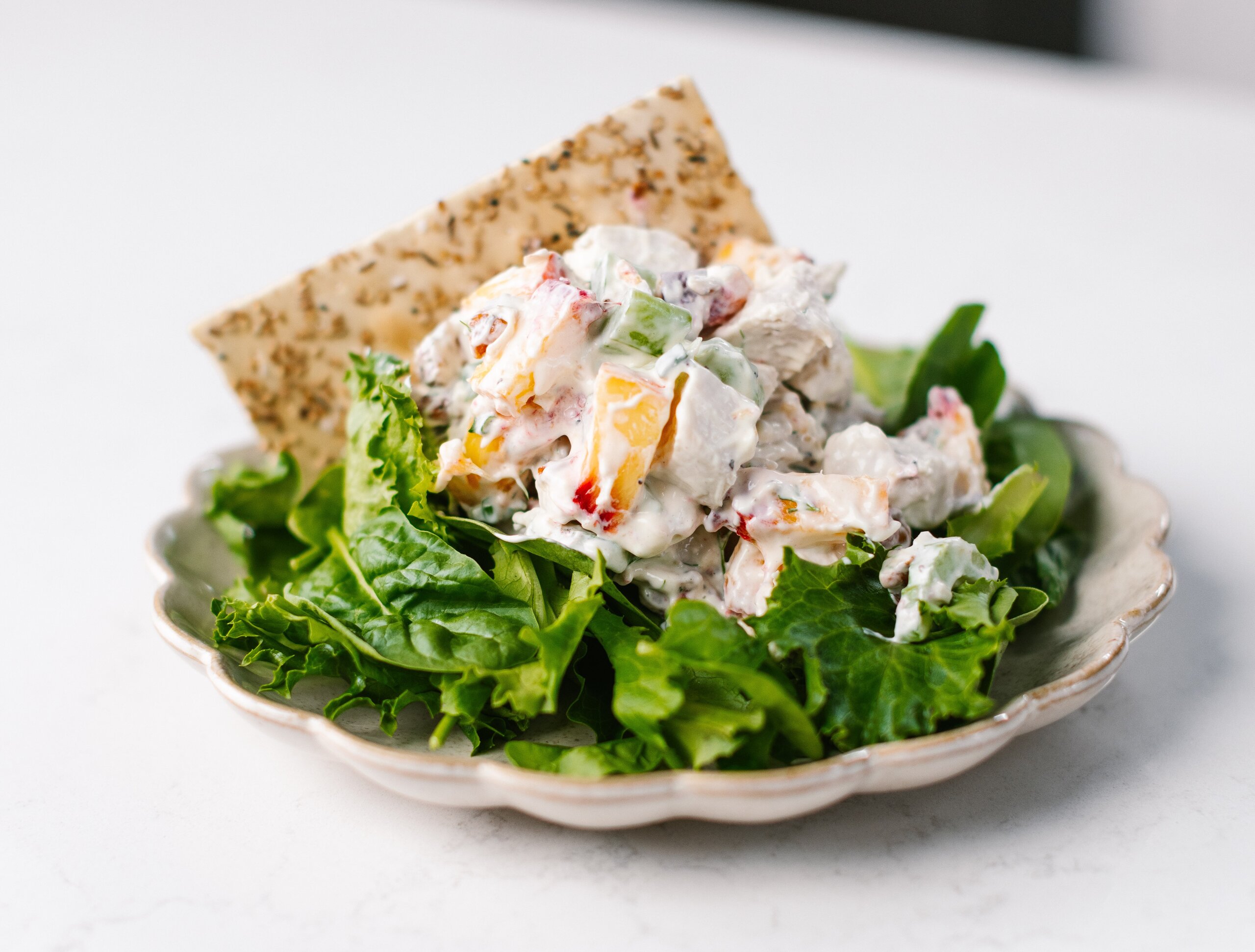 A plate of leafy greens topped with a creamy peach chicken salad containing diced vegetables, served with a piece of seasoned flatbread.