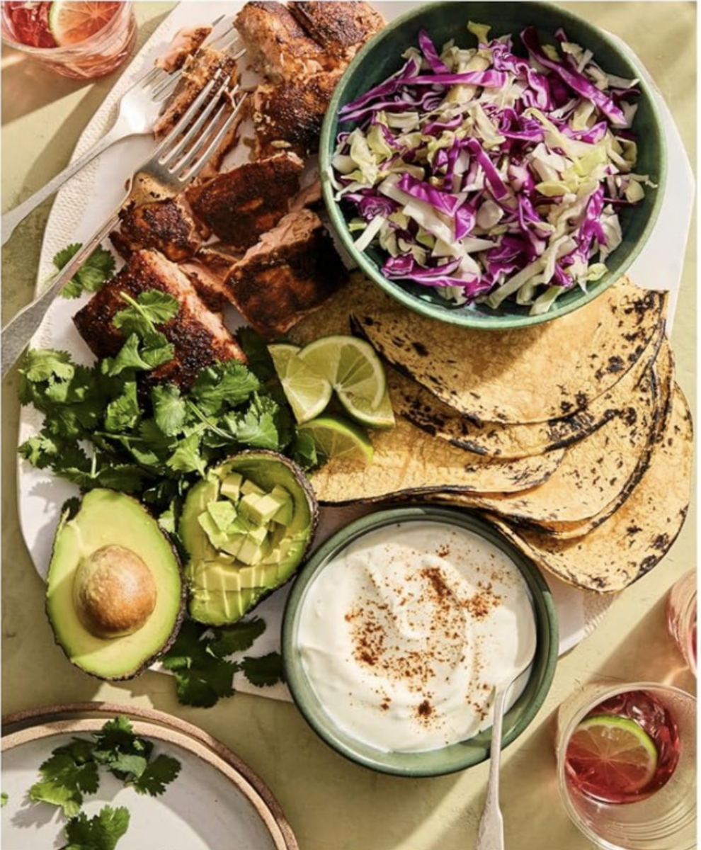 A plate with grilled fish, shredded cabbage, corn tortillas, sliced avocado, lime wedges, fresh cilantro, and a bowl of seasoned sour cream. Drinks are visible in the background.