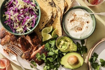 A plate with grilled fish, corn tortillas, shredded cabbage, lime wedges, avocado halves, chopped cilantro, and a bowl of creamy sauce, arranged for a fresh meal.