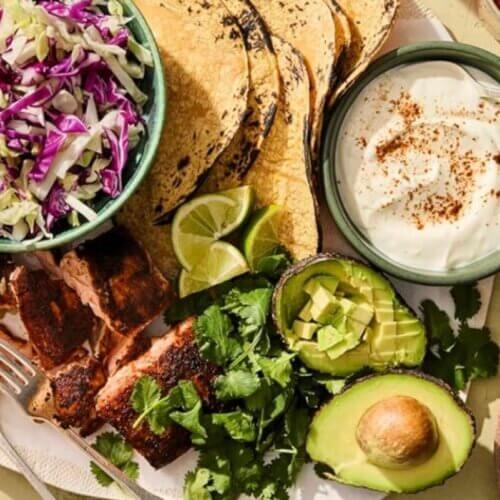 RECIPE: Salmon Tacos - 3 A plate with grilled fish, corn tortillas, shredded cabbage, lime wedges, avocado halves, chopped cilantro, and a bowl of creamy sauce, arranged for a fresh meal.