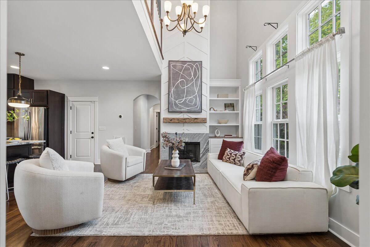 Modern living room with white furniture, large windows, a fireplace, abstract art, and a chandelier. Located at 3014 Forest Wander Way in Nashville, this space features hardwood floors and neutral tones that create a bright, open atmosphere.