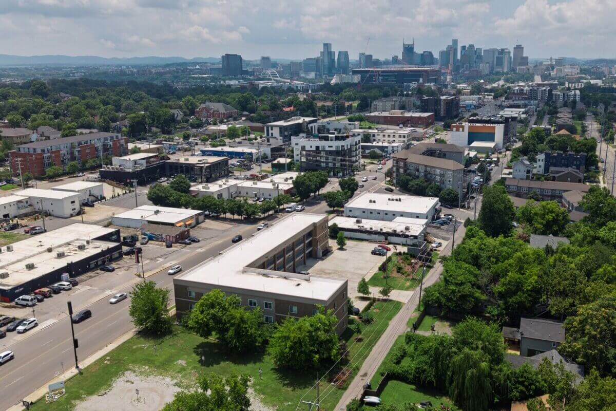 Aerial view of a cityscape with a mix of low-rise and mid-rise buildings, trees, roads, and a distant skyline under a partly cloudy sky, highlighting the vibrant area near 941 Main St Nashville. Explore Nashville TN real estate opportunities like Unit 105 & 106 nestled in this dynamic urban landscape.