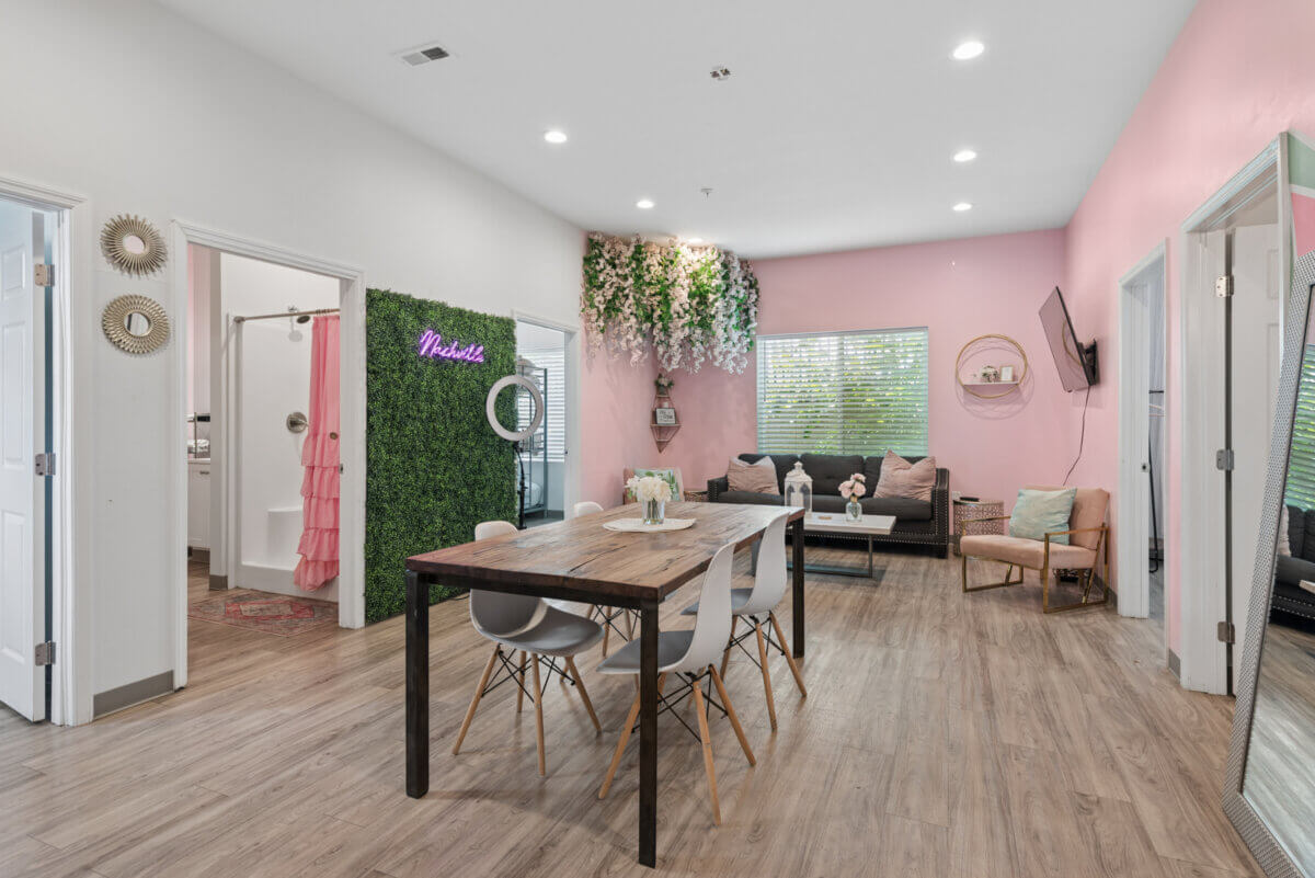 Modern lounge area with a wooden dining table, white chairs, sofa, pink accent wall, greenery decor, and a neon sign that reads "Melanin." Located at 941 Main St Nashville, Unit 106. Open bathroom door is visible on the left.