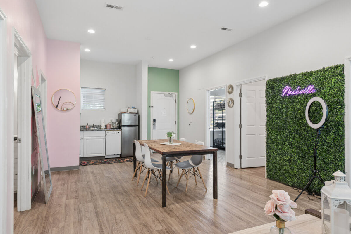 Modern dining area with wooden table, white chairs, kitchenette, green accent wall, neon "Nashville" sign on faux greenery, and a ring light in a bright, well-lit room—perfectly located at 941 Main St Nashville TN, Unit 105 & 106.