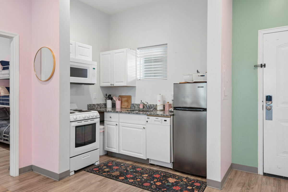 Charming and compact kitchen in Unit 105 at 941 Main St, Nashville, featuring white cabinets, granite countertop, stove, microwave, dishwasher, and a stainless steel fridge. A window and floral rug add warmth to the space; bunk beds are visible in the adjacent room.