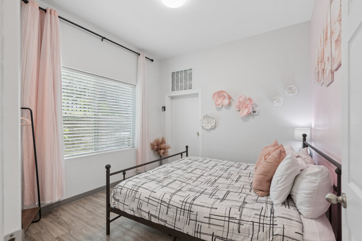 A bedroom in Unit 105 & 106 at 941 Main St Nashville features a black metal bed, patterned bedding, pink accent pillows, wall-mounted flower decorations, a large window with curtains, and light wood flooring—an inviting oasis within sought-after Nashville TN real estate.