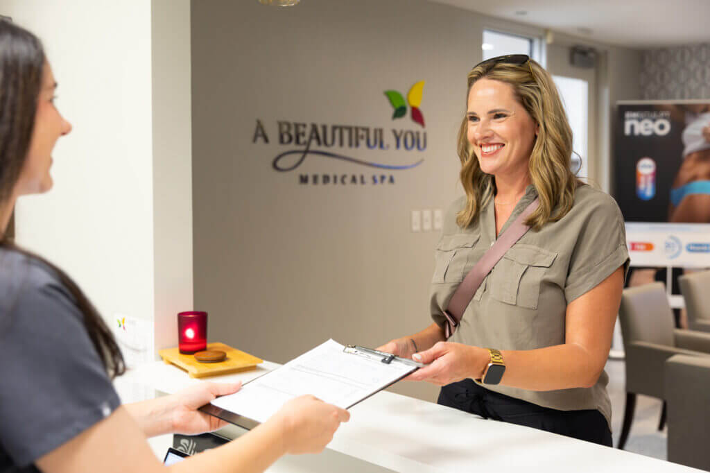 A woman smiles while receiving a clipboard with forms from a receptionist at the front desk of a medical spa called "A Beautiful You," excited to learn more about their innovative peptide therapy offerings.