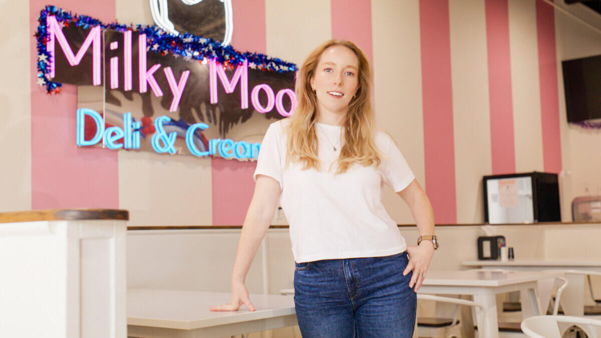 A person in a white shirt and jeans stands in a brightly lit ice cream shop with the "Milky Moo Deli & Creamery" neon sign glowing in the background.