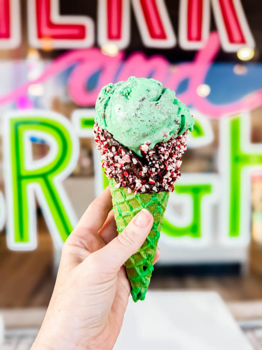 The Inside Scoop on Milky Moo's: Meet the Maker of Alabama's Best Ice Cream - 5 A hand holds a green waffle cone with mint chocolate chip ice cream and a chocolate-dipped rim coated in crushed peppermint, in front of a colorful neon signβcapturing the essence of Alabamaβs best ice cream experience.