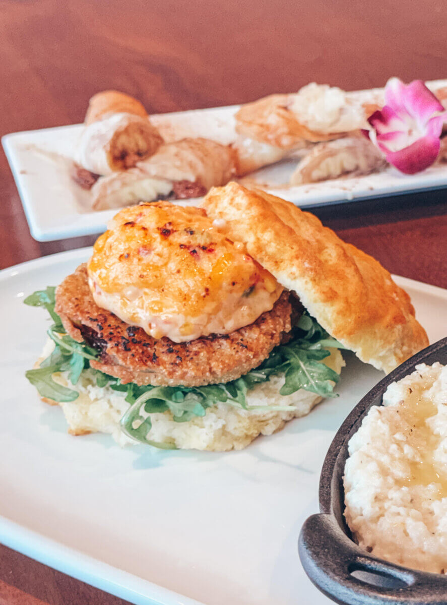 The Inside Scoop on Milky Moo's: Meet the Maker of Alabama's Best Ice Cream - 6 A biscuit sandwich with greens, a fried patty, and sauce, served on a white plate with a side of grits and pastries in the backgroundβbringing a taste of Alabama comfort to your table.