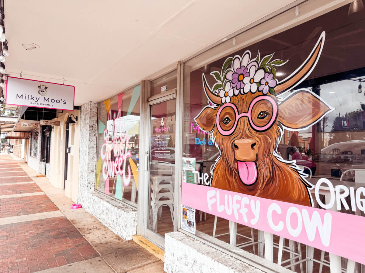 The Inside Scoop on Milky Moo's: Meet the Maker of Alabama's Best Ice Cream - 3 Storefront with a large window mural of a cartoon cow wearing glasses and flowers, next to a sign reading "Milky Moo's"βhome of the best ice cream in Alabama.