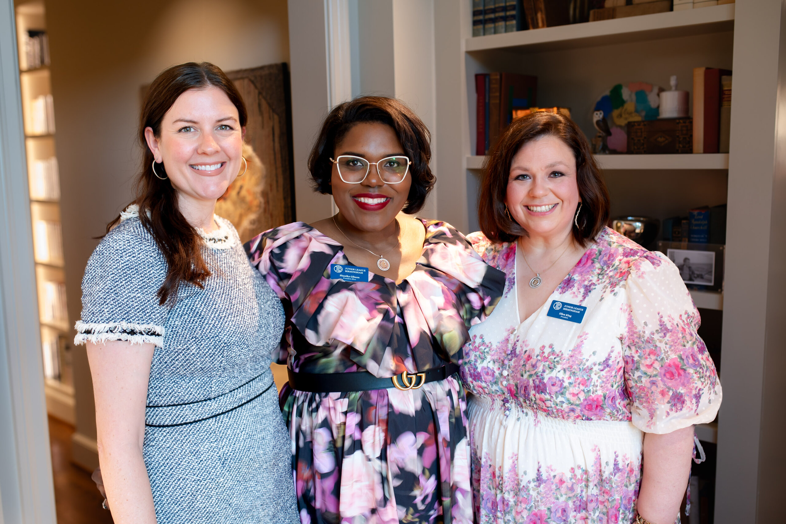 Three women stand together indoors, smiling at the camera. They are wearing patterned dresses and name badges—proudly representing the Junior League of Birmingham—with bookshelves and warm lighting in the background.