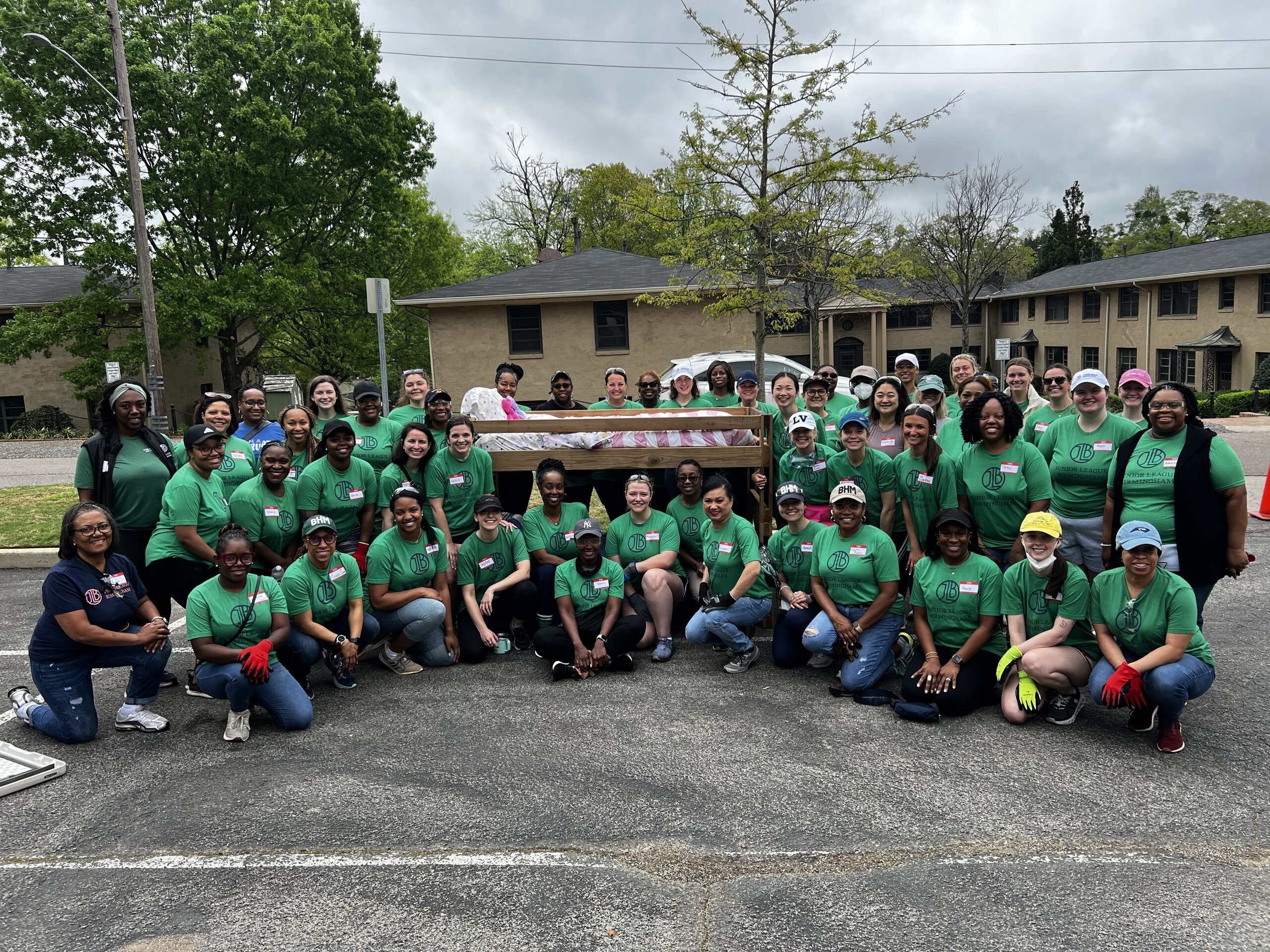 A large group of people in green shirts, representing the Junior League of Birmingham, poses outdoors in front of a truck loaded with bags, with apartment buildings and trees in the background.