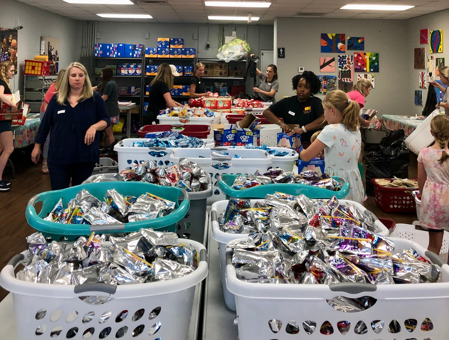People from the Junior League of Birmingham sort and organize food and snacks into laundry baskets in a community room decorated with colorful artwork.