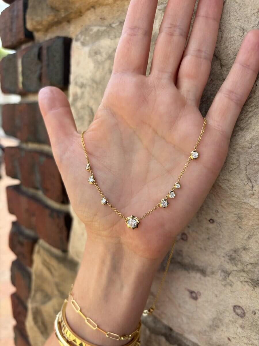 17 Luxurious Birmingham FINDS: Polished Summer Style, Artful Decor & Everyday Luxuries - 2 A hand holds a delicate gold necklace with seven evenly spaced round diamonds—timeless and perfect for everyday luxuries—shown against a stone and brick wall background.