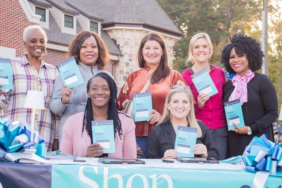 Eight women pose outdoors behind a table, holding up matching books or pamphlets. A banner reading “Shop” is partially visible on the table, suggesting a fundraising or promotional event. The setting appears to be a community gathering organized by the Junior League of Birmingham.
