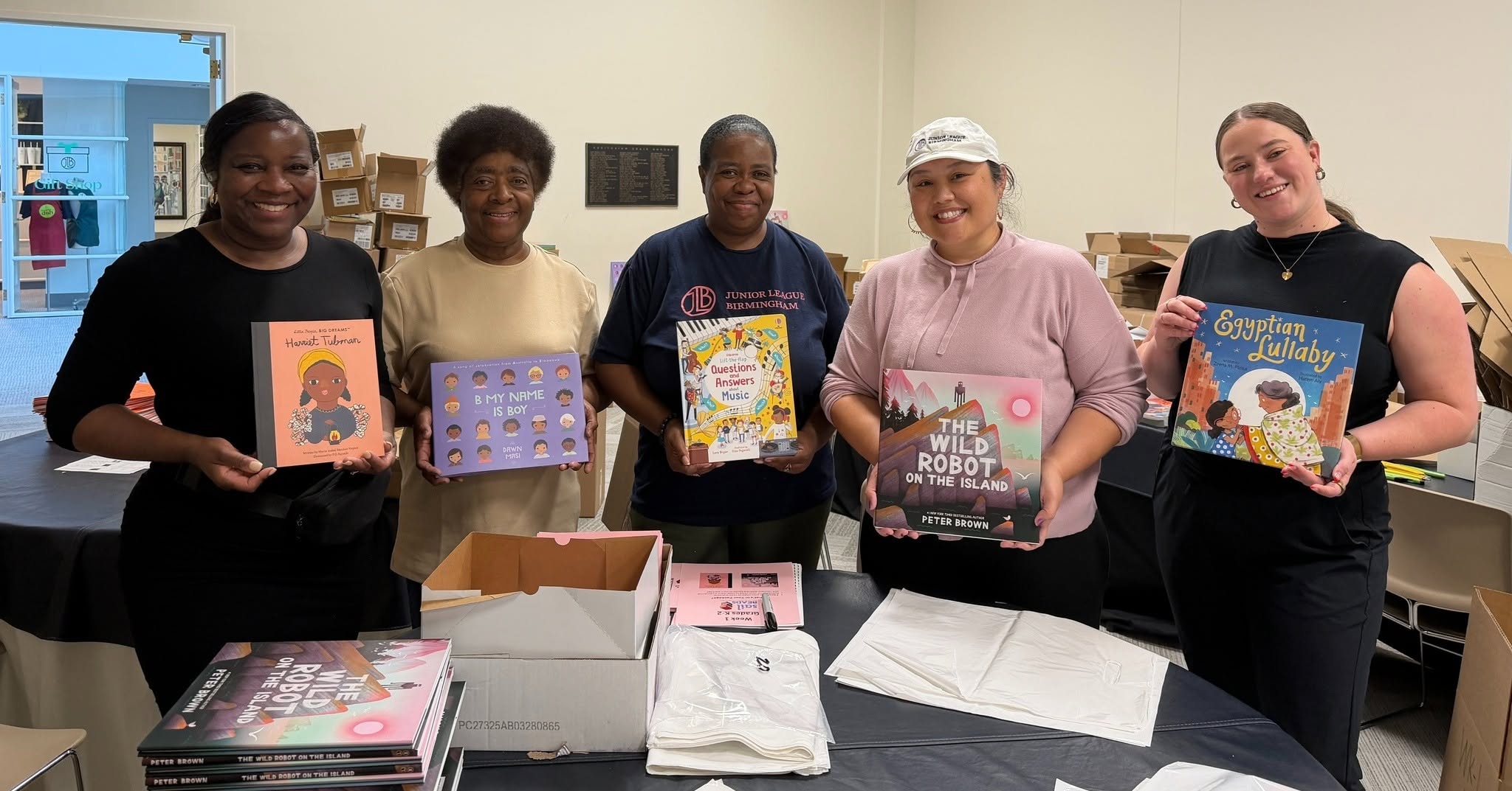 Five people stand in a room holding up children’s books, with more books and white bags on the table in front of them, representing the Junior League of Birmingham’s dedication to promoting literacy in Birmingham.