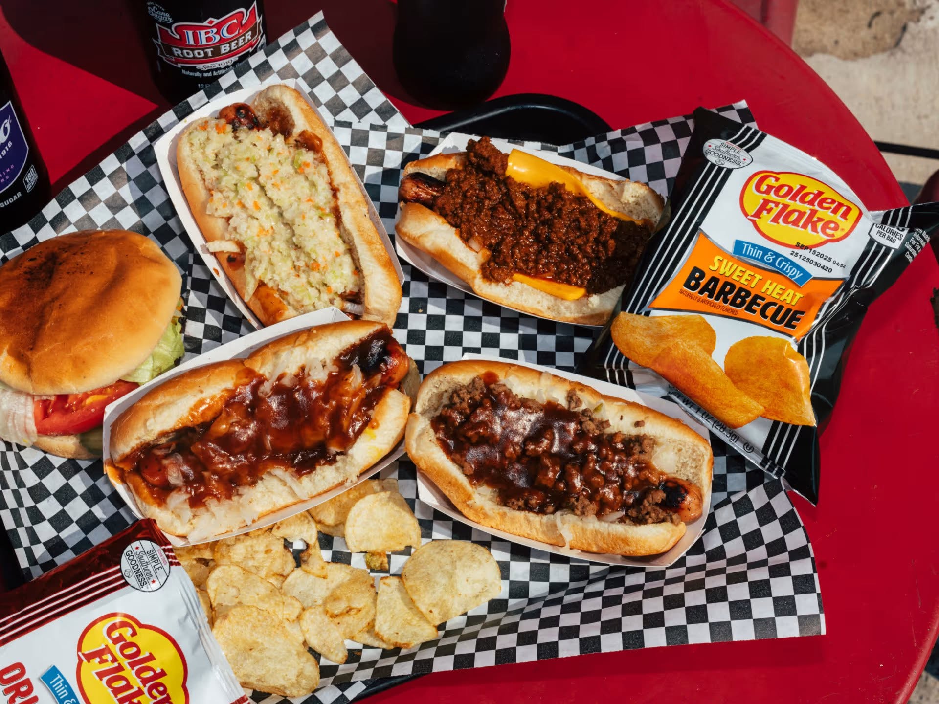 A red table with hot dogs topped with slaw, chili, and barbecue sauce, a hamburger, bags of barbecue chips, and bottles of root beer on a checkered tray—perfect for celebrating Birmingham Restaurant Week and sampling flavors inspired by Birmingham restaurants.